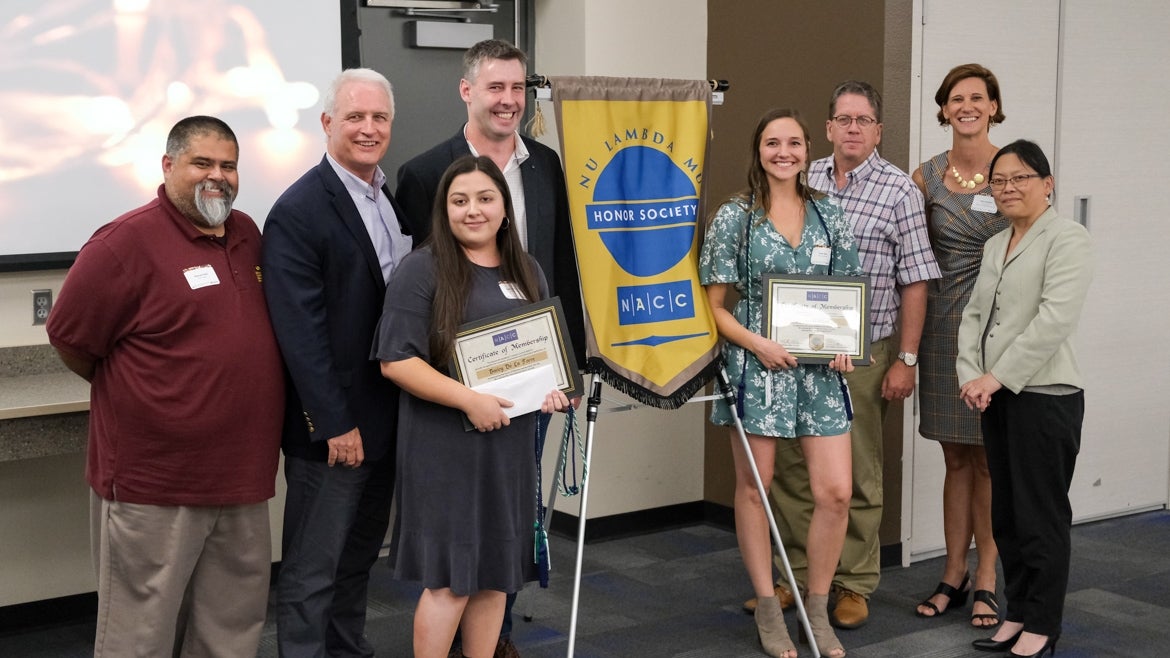 2 undergraduate members of the ASU chapter of the Nu Lambda Mu Nonprofit Honor Society stand and hold certificates. Next to them are ASU faculty members stand behind a banner reading "Nu Lambda Mu, Honor Society, NACC".