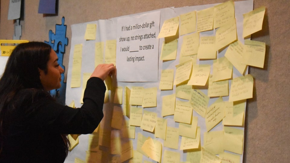 A person puts a yellow post-it note on a wall, next to several other post-it notes. The wall has a large white paper with the words "If I had a million dollars show up, no strings attached, I would (blank) to create a lasting impact."