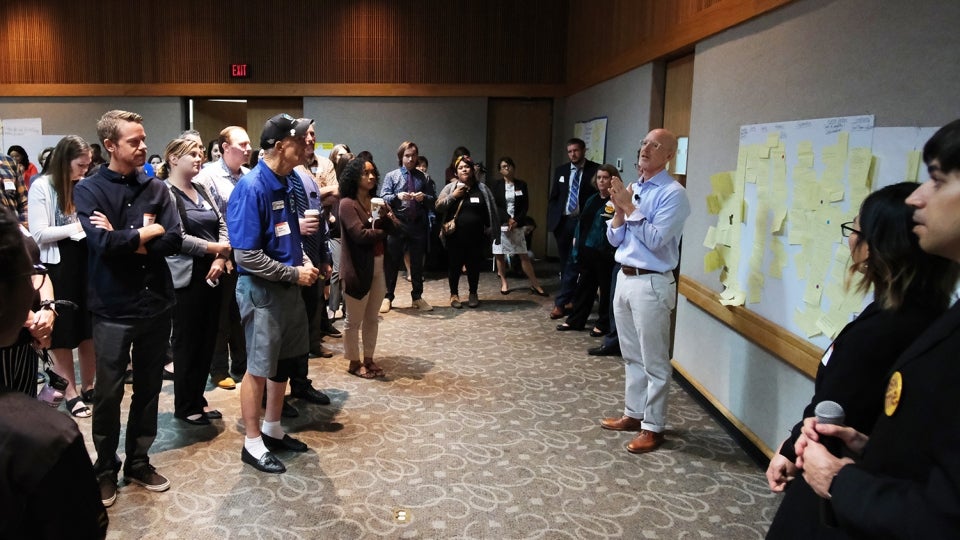 Inside a large room, a person gives a lecture in front of a wall filled with yellow post-it notes. Several people stand and listen.