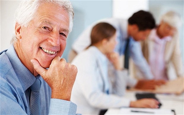 A senior sits and smiles at the camera while others in the background sit at a long table and discuss.