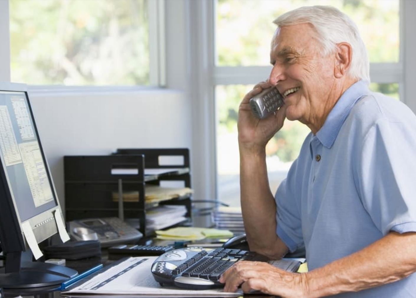 A senior citizen sits at a desk and smiles while on a phone call.