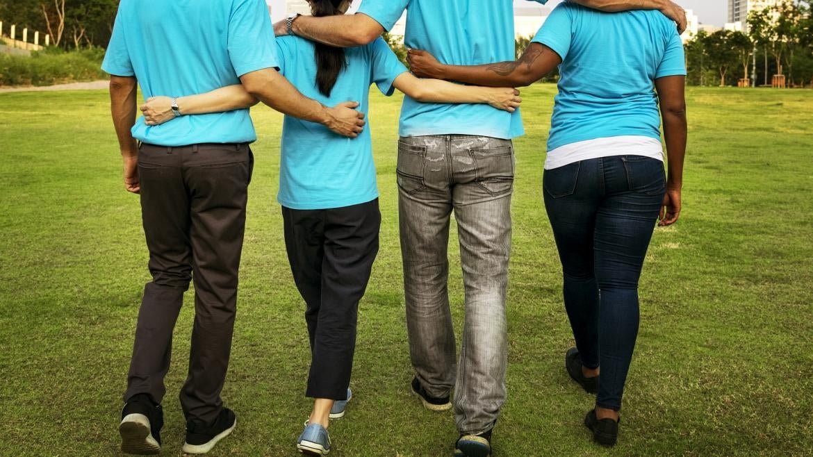 Seen from behind, four people walk across an outdoor grass field with their arms across each others' backs and shoulders.