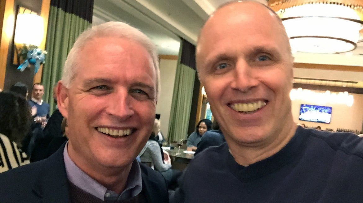 Close up photo of Bill Roske, right, with ASU Lodestar Center Executive Director Dr. Robert Ashcraft. They are inside a room with a TV, table, and crowd in the background.