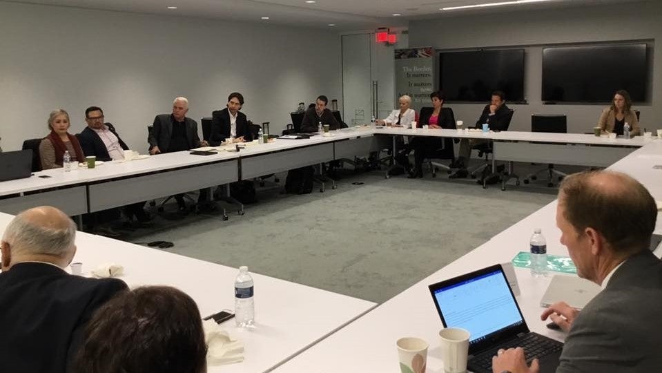 Several people sit in a conference room. There are several tables arranged in a large rectangular so all attendees can see each other.