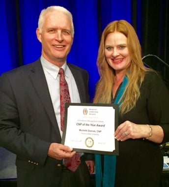 In front of a blue curtain, Michelle Dueñas and Robert Ashcraft stand with a paper certificate.