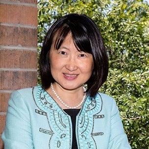 Elena Zee, President & CEO of the Arizona Council on Economic Education and a 2015 alumna of the American Express Leadership Academy at the ASU Lodestar Center, smiles while outside. Behind her is the edge of a brick bulding and plants.