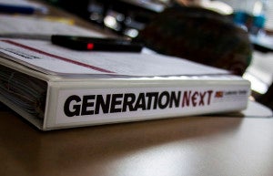 A binder lies on a table. The binder spine says "Generation Next"