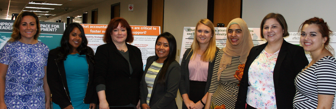 8 of the newest graduates of the ASU Master of Nonprofit Leadership & Management (MNLM) program gather in front of large posters within an office building.