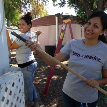 2 people smile at the camera while painting outside. They each hold a long pole with a paintbrush at the end.