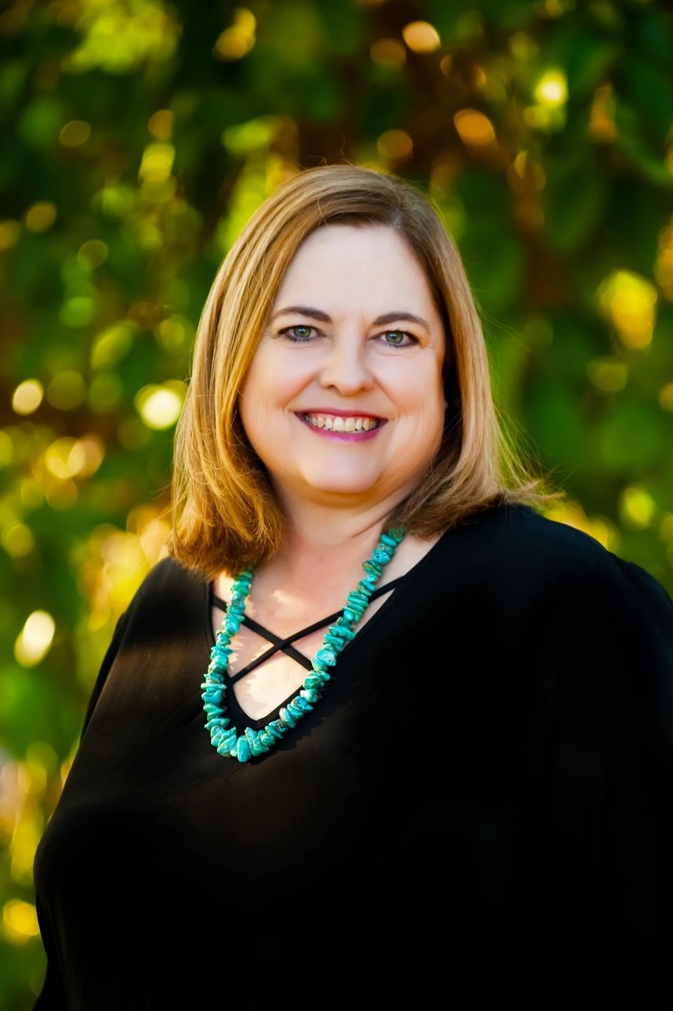 A portrait photo of Rochell Planty outside and smiling. They are wearing a black shirt and turquoise necklace.