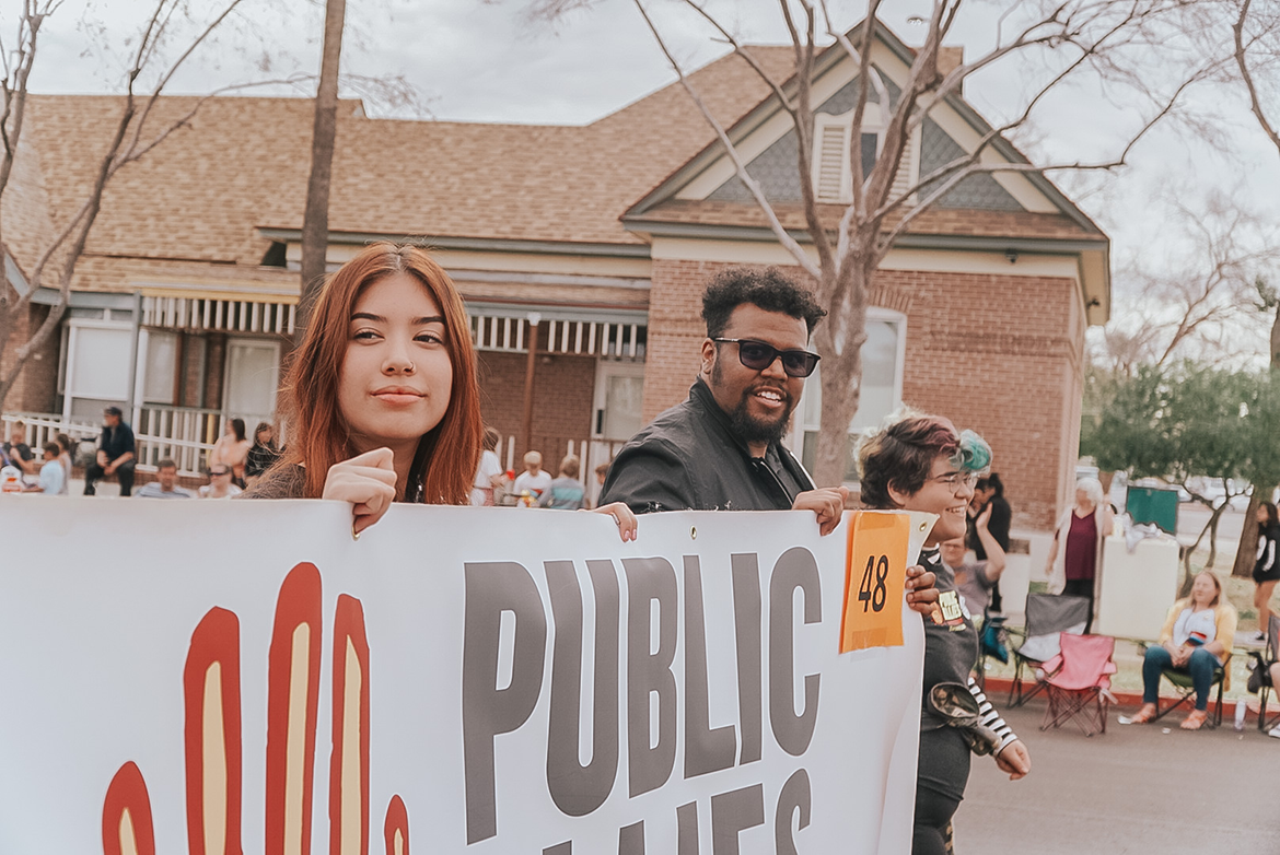 3 people walk down a street while holding a sign displaying the Public Allies logo.