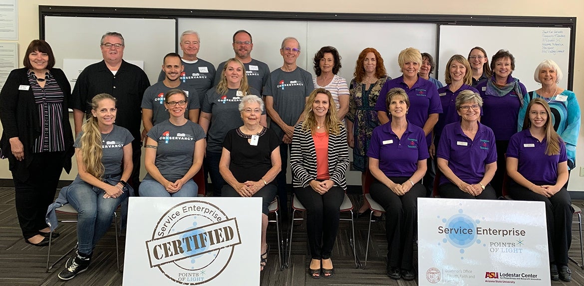 Service Enterprise Initiative Cohort 11 members sit and stand in 3 rows inside a room. Behind them are white boards and in front of them are printed boards reading "Service Enterprise Certified, Points of Light" and "Service Enterprise, Points of Light" alongside logos for ASU Lodestar Center and other organizations.