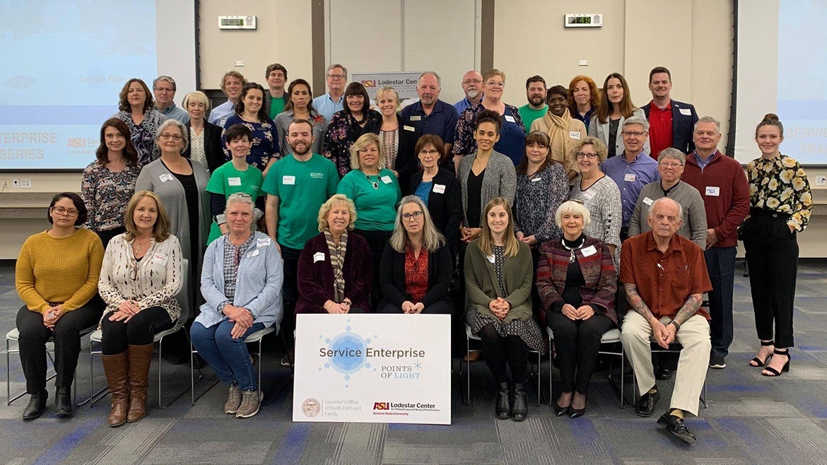 Arizona Service Enterprise Initiative trainees sit and stand in 5 rows within a large room. In the background are projection screens and in front of them is a sign reading "Service Enterprise, Points of Light" with the ASU Lodestar Center logo.