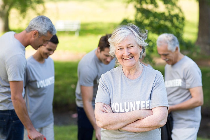 Standing outdoor in a green area, several senior volunteers are busy while one crosses her arms and smiles at the camera.