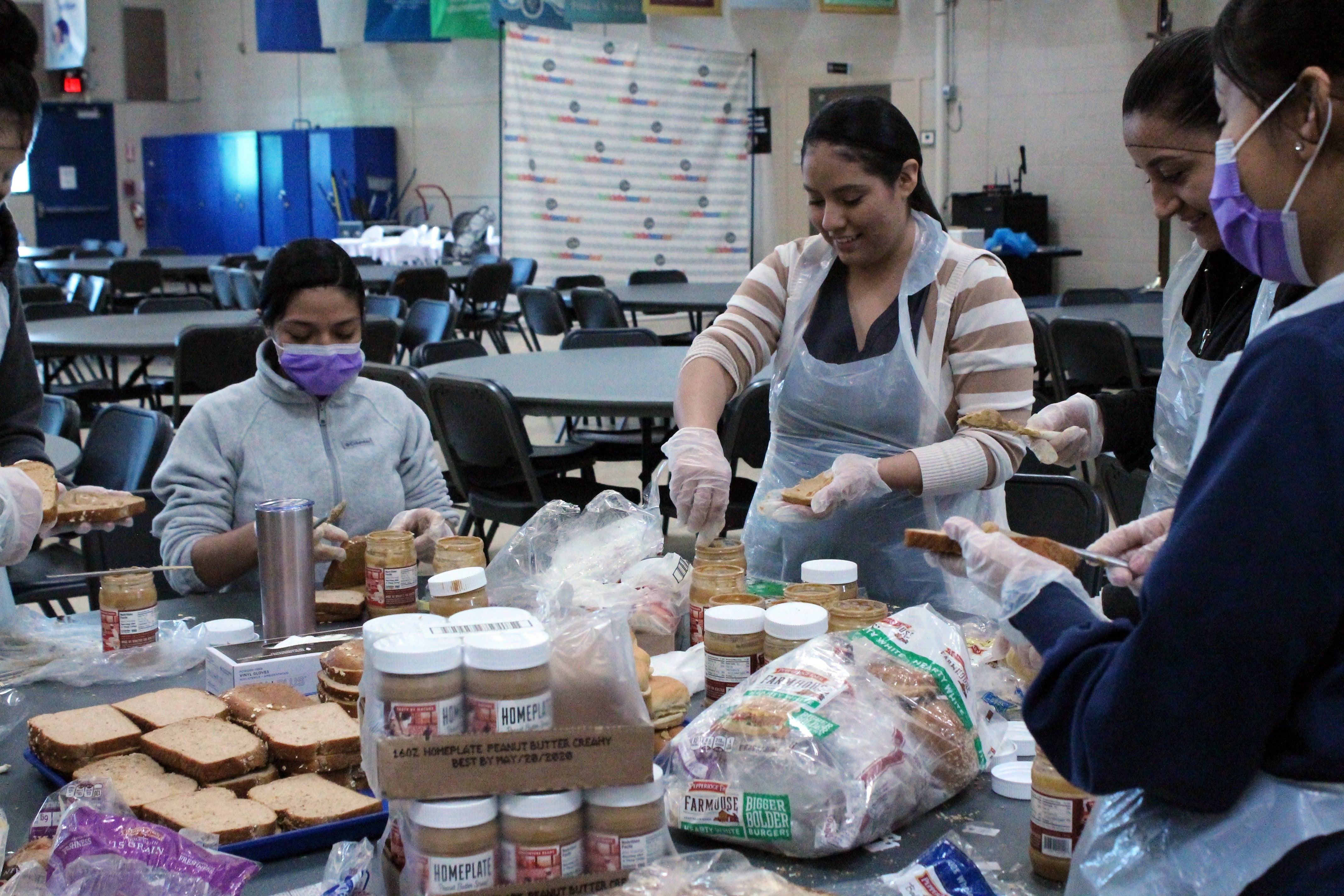 Several people stand around a table filled with food items. They are making sandwiches.