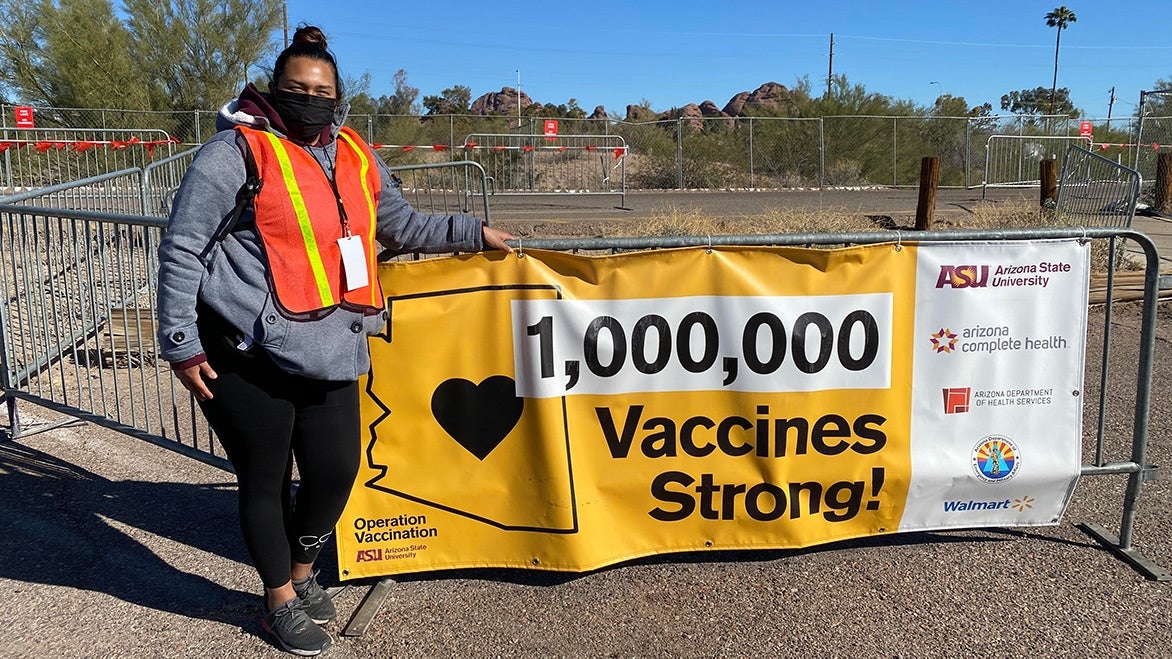 A person wearing a face mask and bright orange vest poses by a sign reading "1,000,000 vaccines strong!".